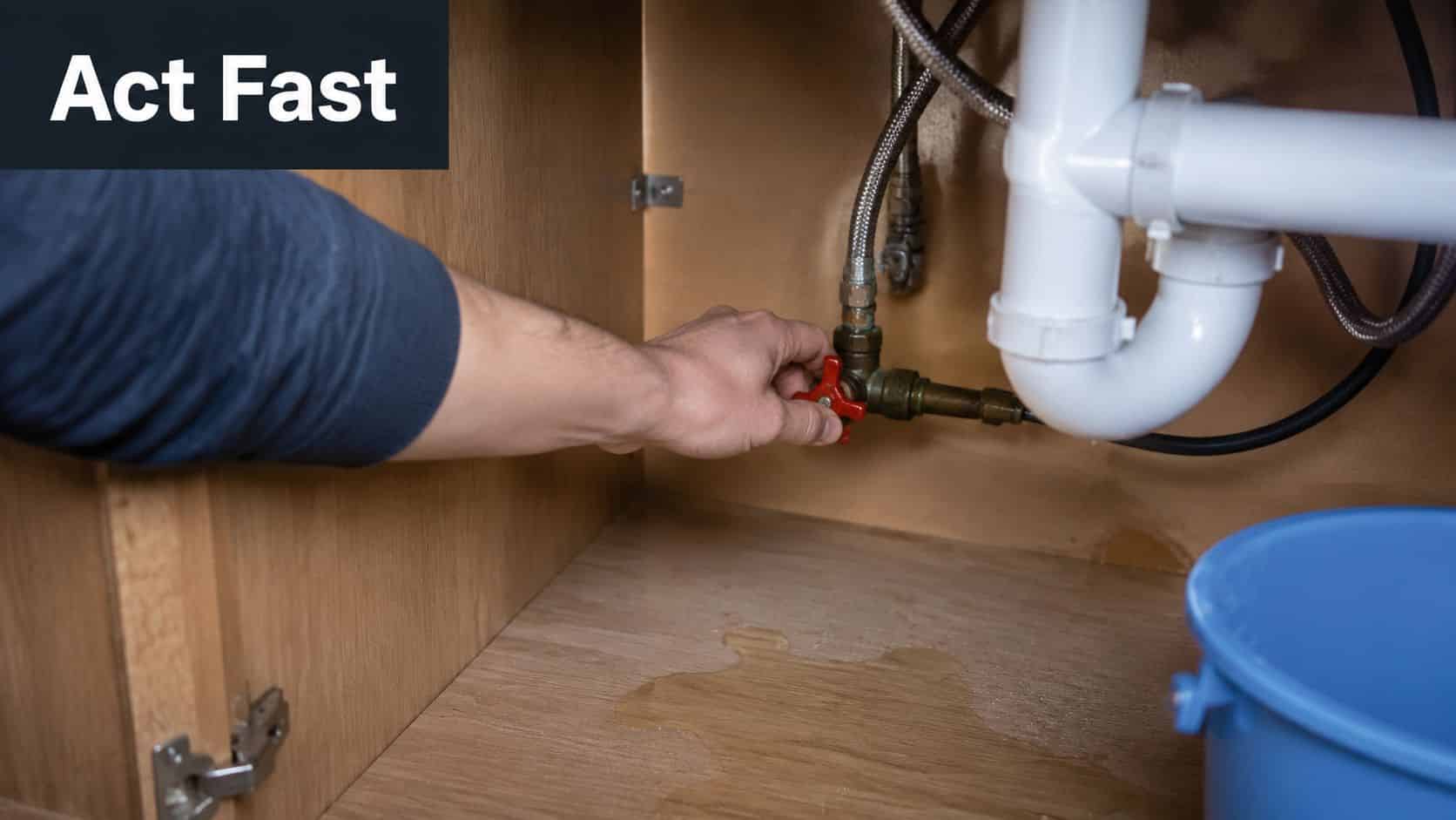 A person turning a red shut-off valve under a kitchen sink to stop a water leak.